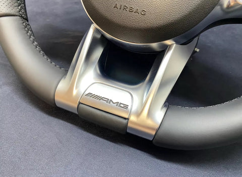 A steering wheel with "AMG" branding, featuring leather and metallic detailing. The word "AIRBAG" is embossed on the central section, set against a dark background.