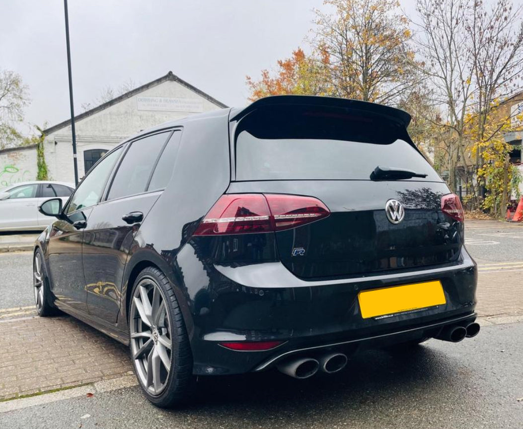 A black hatchback car is parked on a street near autumn trees and a warehouse. It features dual exhausts and alloy wheels, with a visible "VW" logo on the rear.