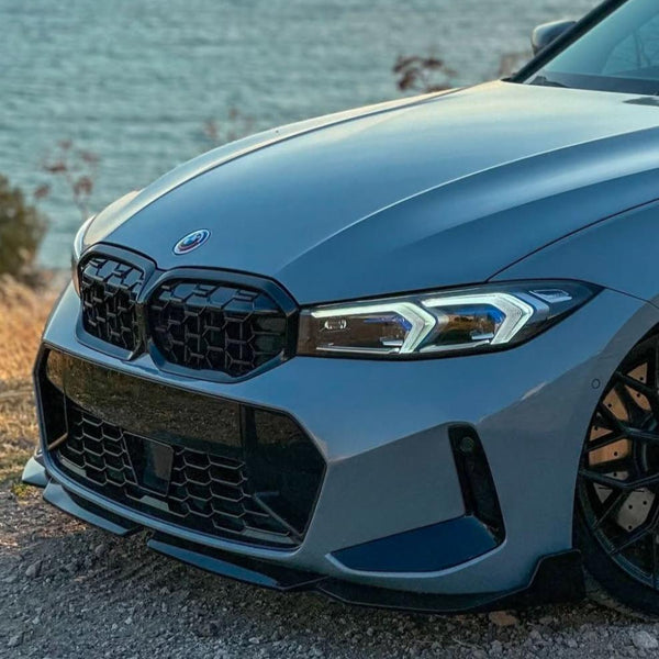 A blue car with a sleek front grille and sharp headlights is parked on a rocky terrain, overlooking a large body of water in the background.