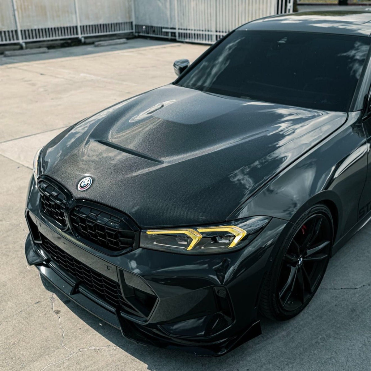 A sleek, dark-colored car with tinted windows and yellow headlights is parked on a concrete surface, surrounded by a metal fence. The car features a prominent front grille.
