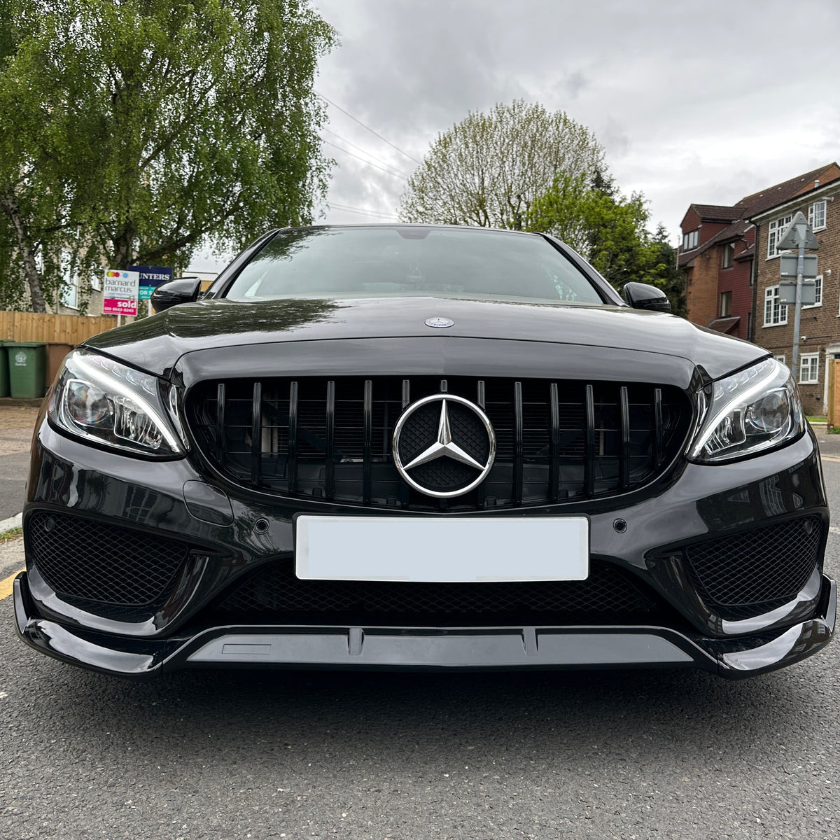 Black Mercedes car parked on a residential street with trees, brick buildings, and several trash bins in the background. A "For Sale" sign is visible among the greenery.