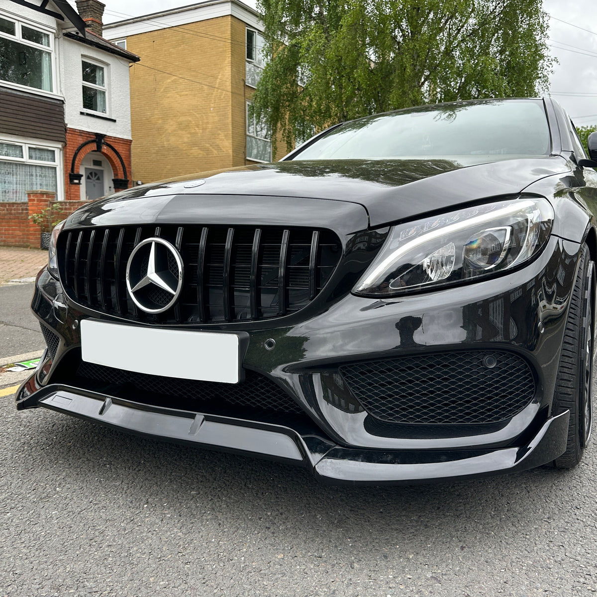 Car parked on a residential street, featuring a glossy black finish and a prominent logo on the front grille. Surrounded by brick and cream houses with visible driveway and greenery.