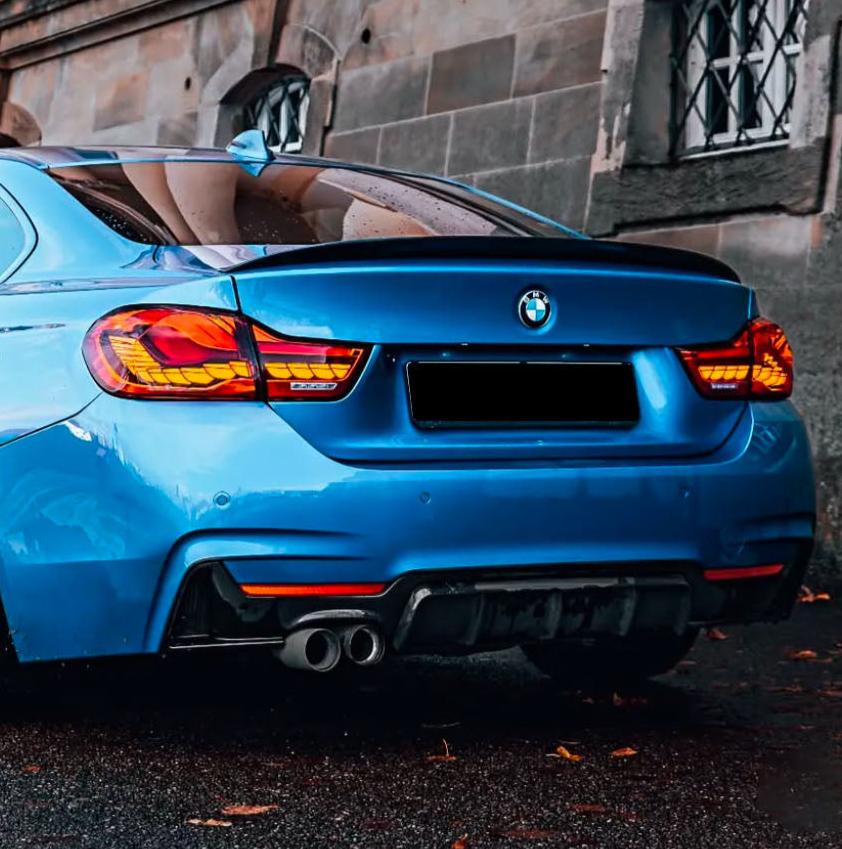 A blue BMW car is parked, showing its rear with dual exhaust pipes. It's situated beside a stone building with barred windows, on a wet pavement scattered with leaves.
