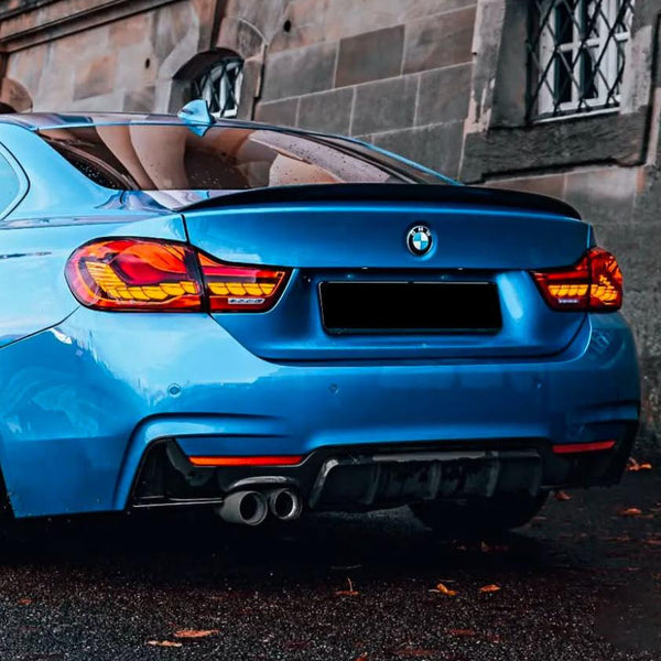 A blue BMW car is parked, showing its rear with dual exhaust pipes. It's situated beside a stone building with barred windows, on a wet pavement scattered with leaves.