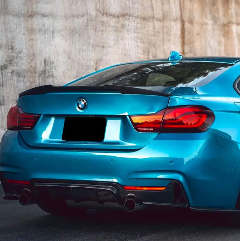 A blue BMW car is parked beside a concrete wall, showing its rear end with dual exhaust pipes and the BMW logo centered on the trunk.