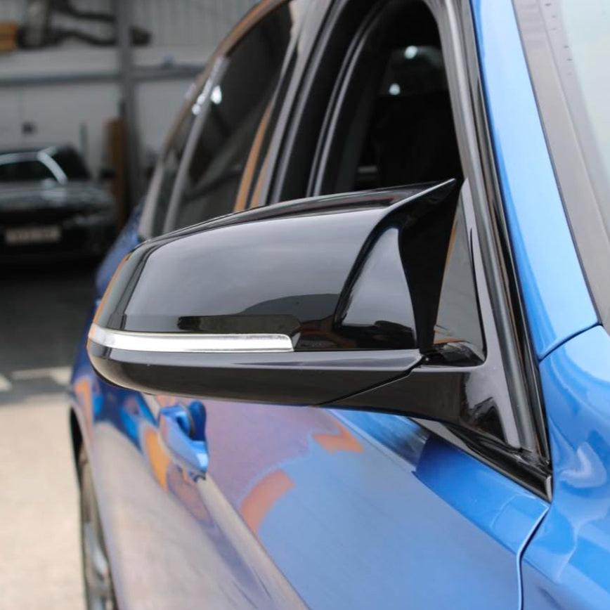A sleek black side mirror is attached to a blue car, reflecting part of a garage interior, with cars and industrial elements in the background.