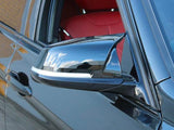 A black car's side mirror reflects blue sky with clouds. The car has red leather interior seats, parked against a brick wall in sunlight.