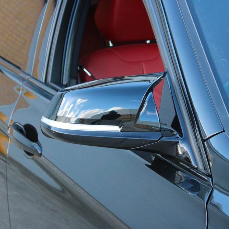 A black car's side mirror reflects blue sky with clouds. The car has red leather interior seats, parked against a brick wall in sunlight.