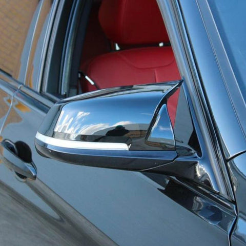 A sleek black car mirror reflects the blue sky and clouds, attached to a vehicle with red interior seats, parked near a brick wall.