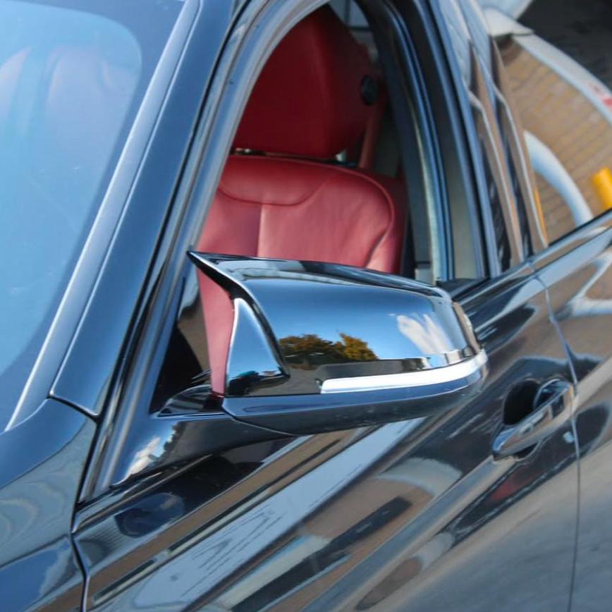A black car's side mirror reflects the surrounding sky and trees. The car’s window is slightly open, revealing red leather seats inside. The setting includes a cityscape with buildings.