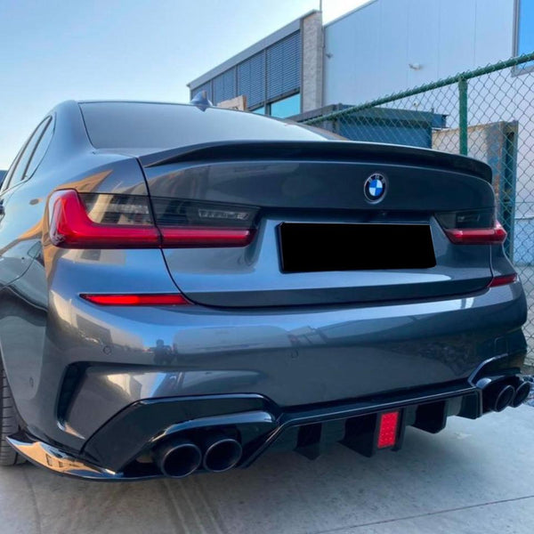 A dark BMW car parked near a fencing; its distinctive rear lights and dual exhausts stand out. The background includes a modern building under a clear sky.