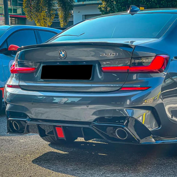 A gray BMW 330i car is parked, with red and black accents, alongside a blue vehicle. The location is a sunny outdoor lot near buildings and palm trees.