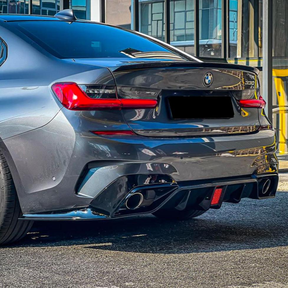 A silver BMW car with sleek taillights and dual exhaust is parked on an asphalt surface, reflecting nearby buildings. The model name "330i" is visible on the trunk.
