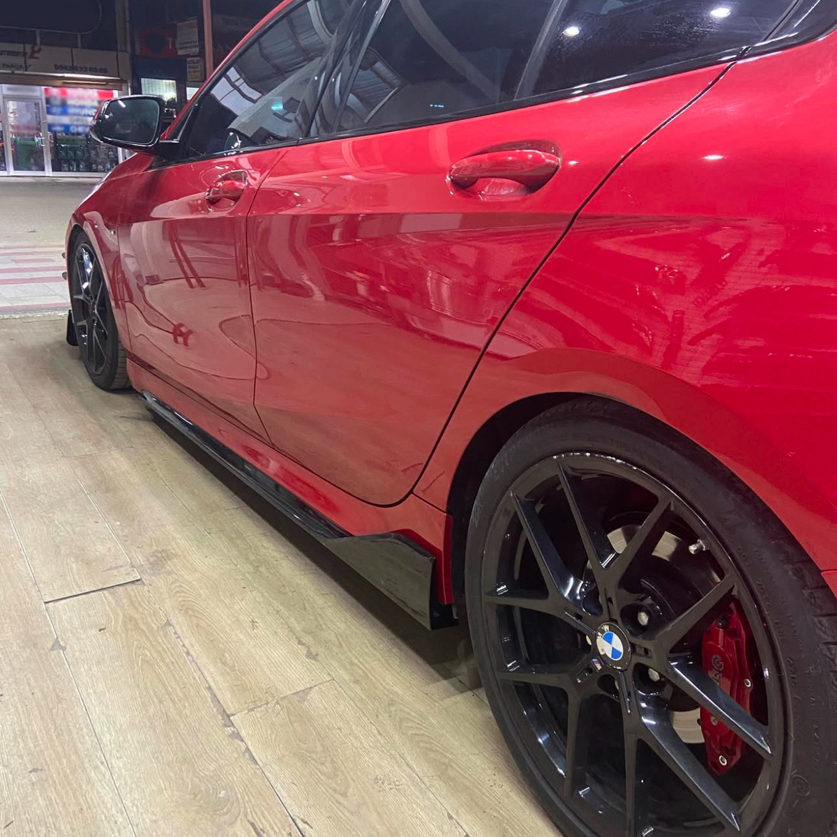 A red car with black rims is parked indoors on a wooden floor, reflecting overhead lights and nearby structures in a garage setting.