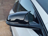 A black side mirror reflects a clear sky, attached to a white car. The vehicle is parked on a gravel surface, with a wooden fence in the background.