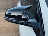 A sleek black side mirror is attached to a white car, reflecting a blurred outdoor scene, with a gravel ground visible in the background.