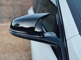A sleek black car side mirror reflects surroundings, attached to a white vehicle parked outdoors on a gravel surface with a blurred wooden backdrop.