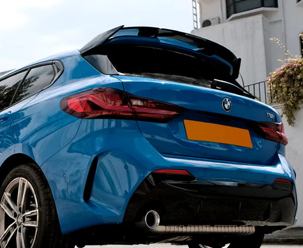 A blue BMW car is parked, showcasing its rear with a visible exhaust pipe and taillights. It's positioned near a white building with a small, flowering plant.