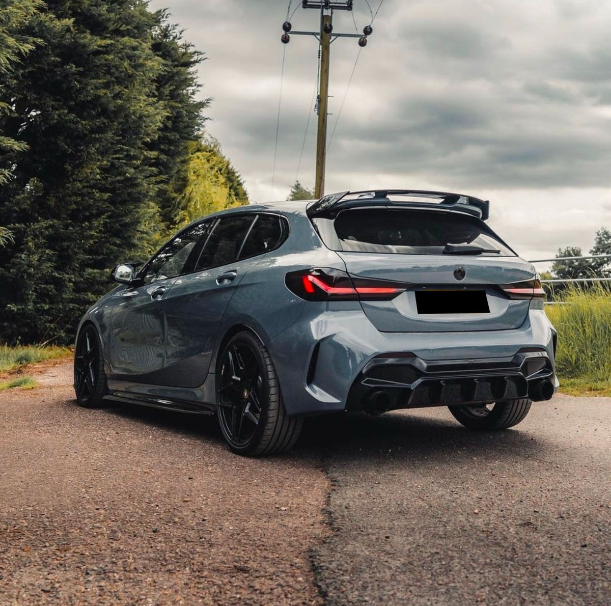 A sleek blue car is parked on a paved road, surrounded by lush green trees and under a cloudy sky. The car features tinted windows and a prominent rear spoiler.