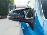 A black side mirror is attached to a blue car, reflecting the sky and surrounding greenery; the scene is outdoors in a paved area with a fence in the background.
