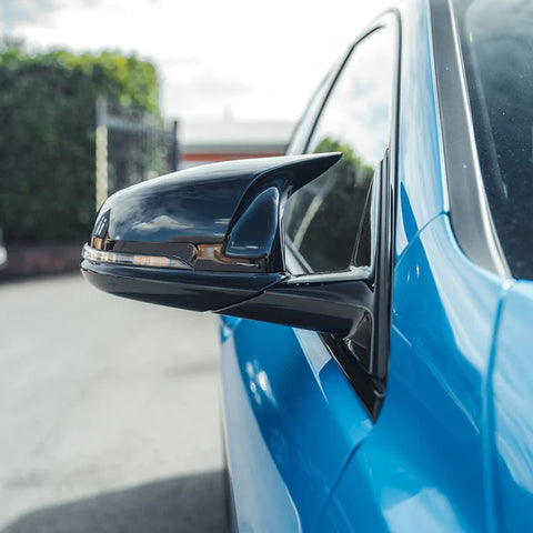 A black side mirror is attached to a blue car, reflecting the sky and surrounding greenery; the scene is outdoors in a paved area with a fence in the background.