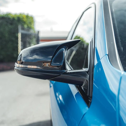 A car's sleek, black side mirror reflects its surroundings, attached to a bright blue vehicle, parked on a sunlit street with trees and buildings in the distant background.
