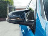 A shiny black side mirror is attached to a bright blue car, reflecting a cloudy sky. The vehicle is parked near a hedge and gate under a partly cloudy sky.