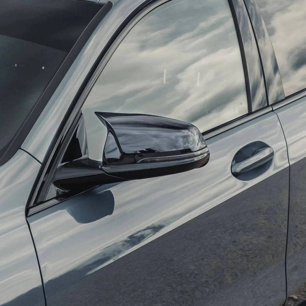 The side of a sleek car features a shiny side mirror reflecting clouds. The car's metallic surface also shows a mirror-like reflection, set outdoors under a bright sky.