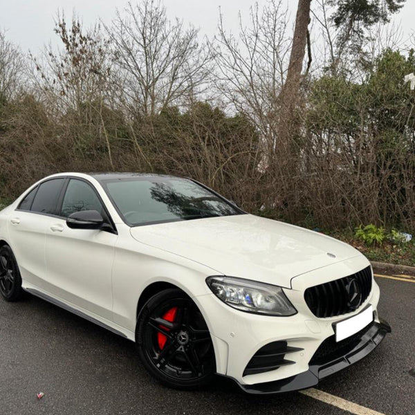 A white car with black rims is parked on a wet road beside leafless trees and bushes. The vehicle features a sleek design with red brake calipers.