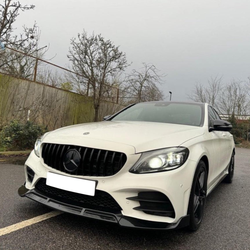 A white car with illuminated headlights is parked on a wet pavement, surrounded by leafless trees and a wooden fence under a cloudy sky.