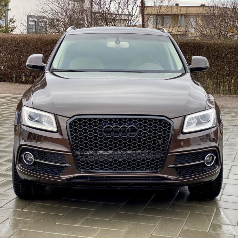 A brown Audi SUV parked on a tiled driveway, surrounded by leafless trees and residential buildings in the background, facing the camera directly.