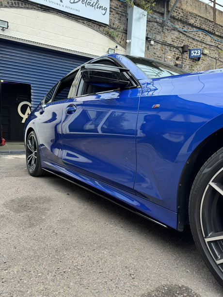 Blue car parked, driver's window partially open, outside a building with a blue garage door. Sign reads “Styling & Detailing.” Number 523 is visible on the brick wall.