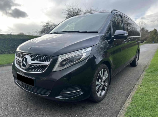 A black Mercedes-Benz van is parked on an asphalt road, bordered by grass and hedges, under a cloudy sky.