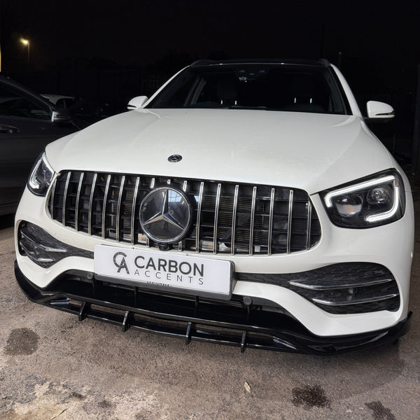 A white luxury car with a prominent emblem and grille, parked in a dimly lit area. The front license plate reads "CARBON ACCENTS."
