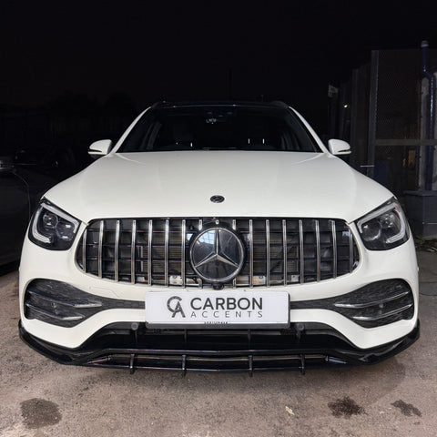 A white SUV with a prominent front grille bears a badge and is parked in a dimly lit area. The license plate reads "CARBON ACCENTS AUTOMOTIVE."