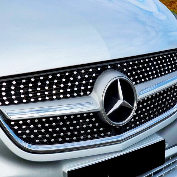 A silver car grille displays a circular emblem with a three-point star, surrounded by a pattern of dots. It's parked on an asphalt road near green grass.
