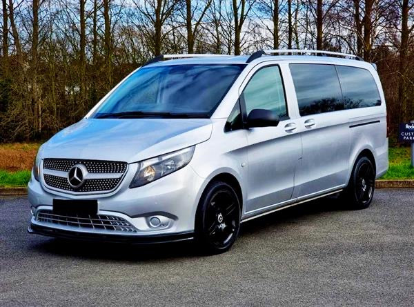 A silver van with a Mercedes-Benz logo is parked on a paved road. It's surrounded by leafless trees, indicating a winter setting.