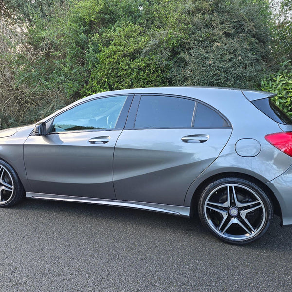 A silver car is parked on a road beside dense green bushes. It features sleek curves, dark-tinted windows, and modern, star-shaped alloy wheels.