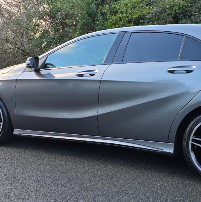 A sleek, gray car is parked on a paved road, with its tinted windows reflecting light. Surrounding context includes lush green foliage under clear skies.