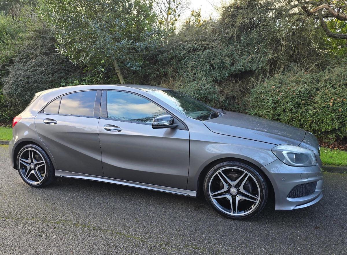A silver hatchback car is parked on a paved road, surrounded by lush, green shrubbery and trees on a clear day.
