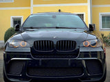 A black BMW with illuminated circular headlights is parked in front of a yellow building with white trim and windows, creating a striking contrast between the car and its surroundings.