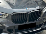 A shiny gray BMW car with a prominent black grille and blue-accented headlights is parked on interlocking paving stones. A building is reflected on the hood.