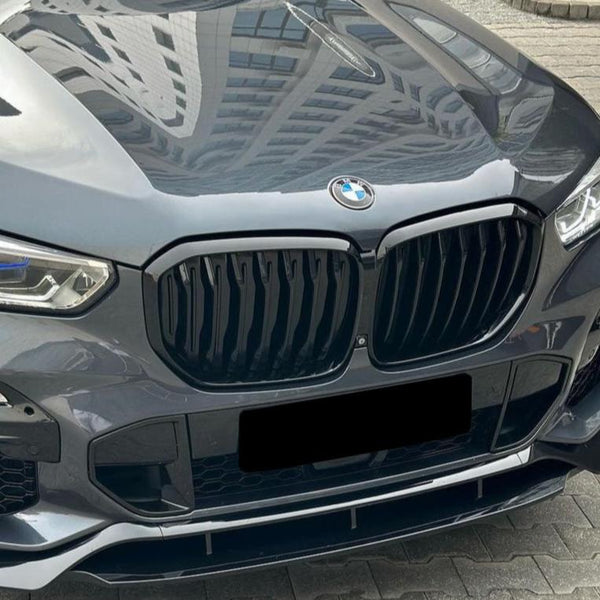 A shiny gray BMW car with a prominent black grille and blue-accented headlights is parked on interlocking paving stones. A building is reflected on the hood.