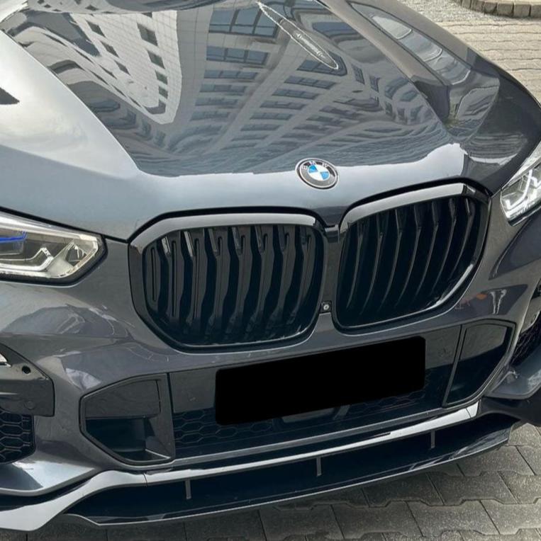 A shiny gray BMW car with a prominent black grille and blue-accented headlights is parked on interlocking paving stones. A building is reflected on the hood.