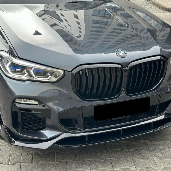 A sleek, gray car with a prominent grille and shiny, intricate headlights is parked on a patterned concrete surface, reflecting nearby buildings on its glossy hood.