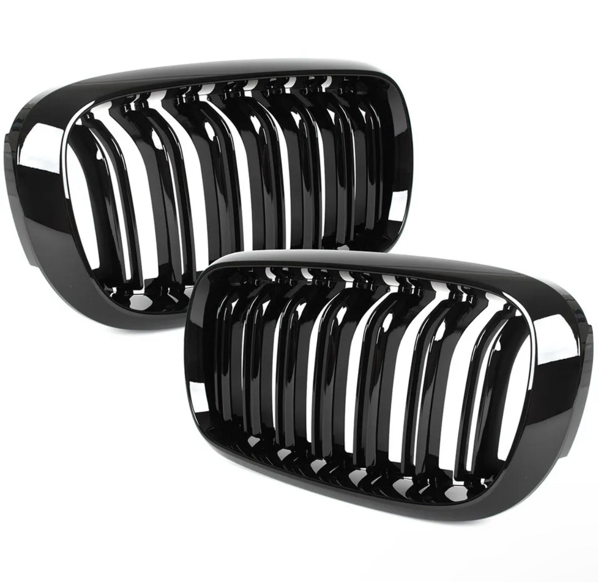 Two black, glossy car grilles with vertical slats lean against each other, reflecting light. They are set against a plain white background.