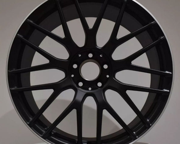A black alloy wheel with multiple spokes, labeled "AMG" at the top, is resting upright against a neutral background atop a white circular platform.
