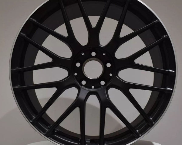 A black AMG car wheel rim with a multi-spoke design is displayed against a plain background, positioned upright on a circular white platform.
