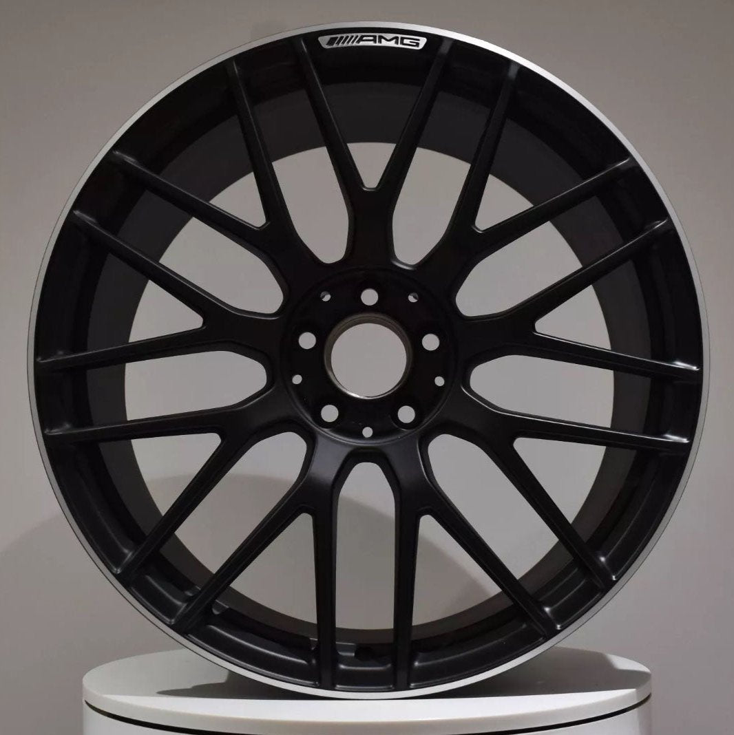 A black car wheel with multiple spokes displays the "AMG" logo at the top, set against a plain gray background on a white surface.