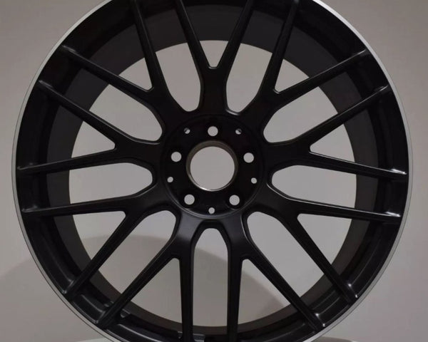 A black, multi-spoke AMG car rim is displayed upright against a neutral background, resting on a white surface.